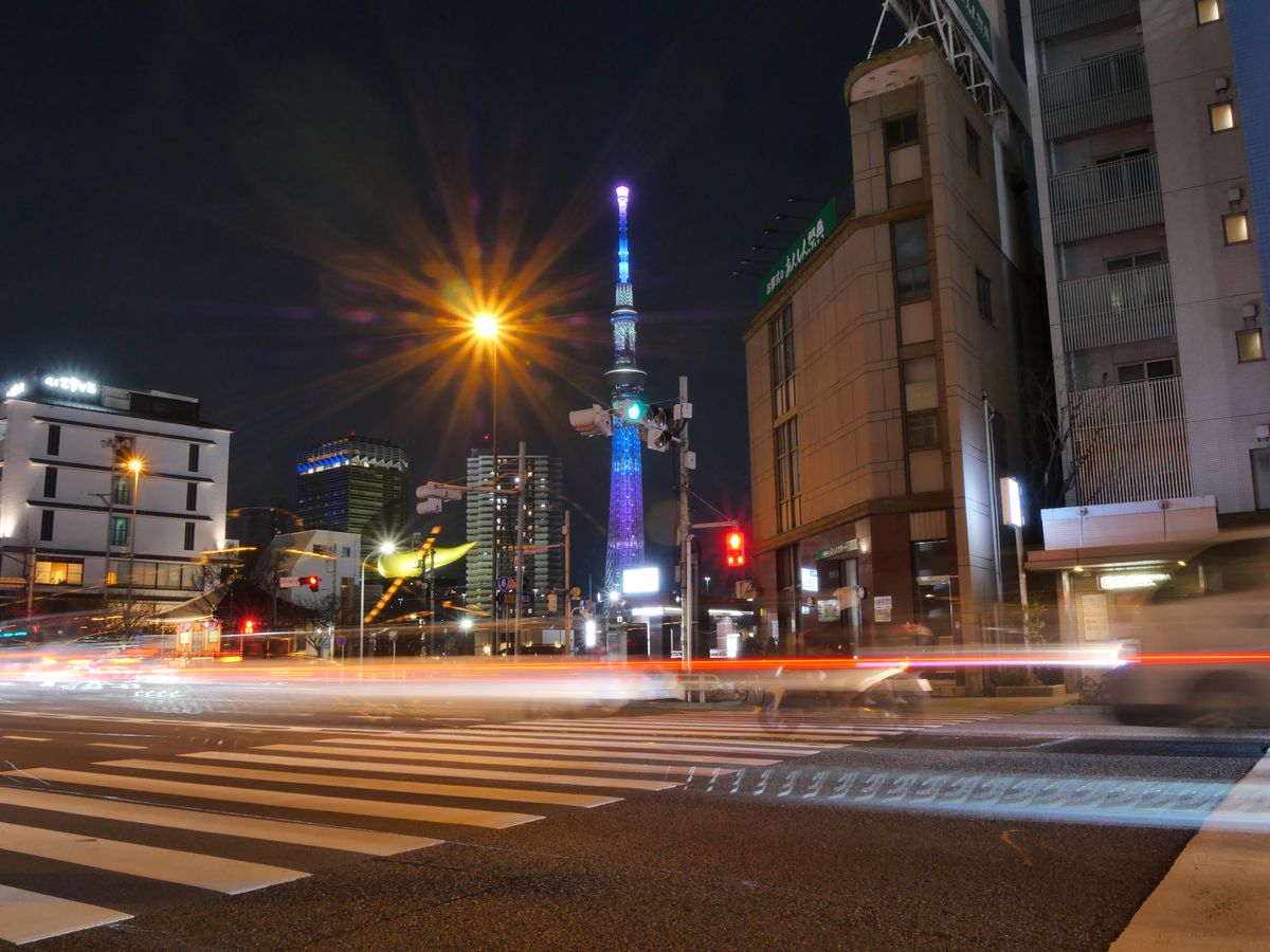 Tokyo Skytree lit purple and blue at night above a crossing streaked with light trails